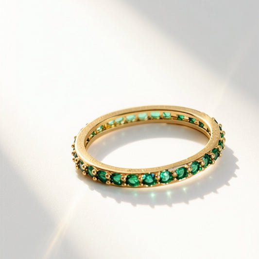A gold eternity ring set with round green emerald-cut gemstones, photographed on a clean white surface with artistic diagonal shadows.