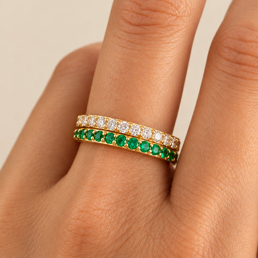 A close-up of a hand wearing two gold eternity rings. One ring is set with clear stones, and the other is set with round green emeralds.