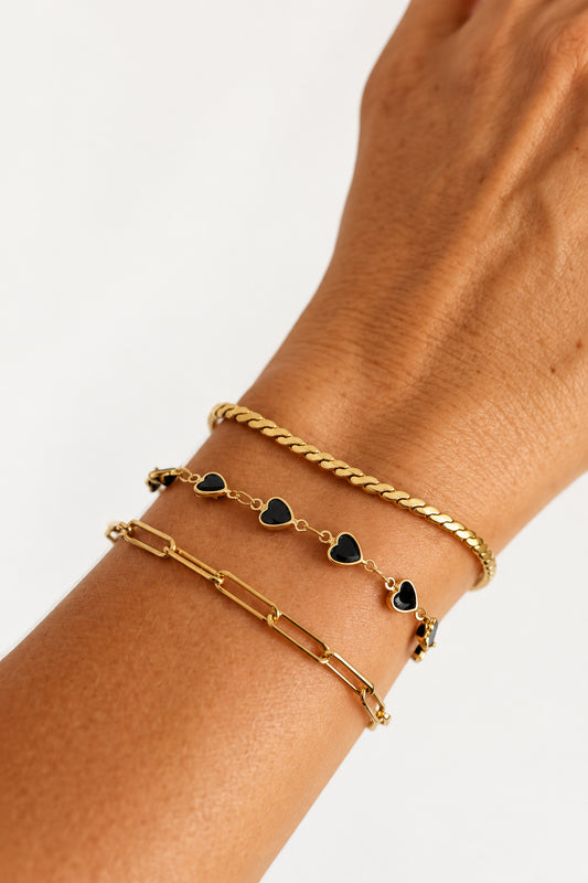 Three gold-toned bracelet with heart-shaped charms, worn on a person's wrist against a light neutral background.