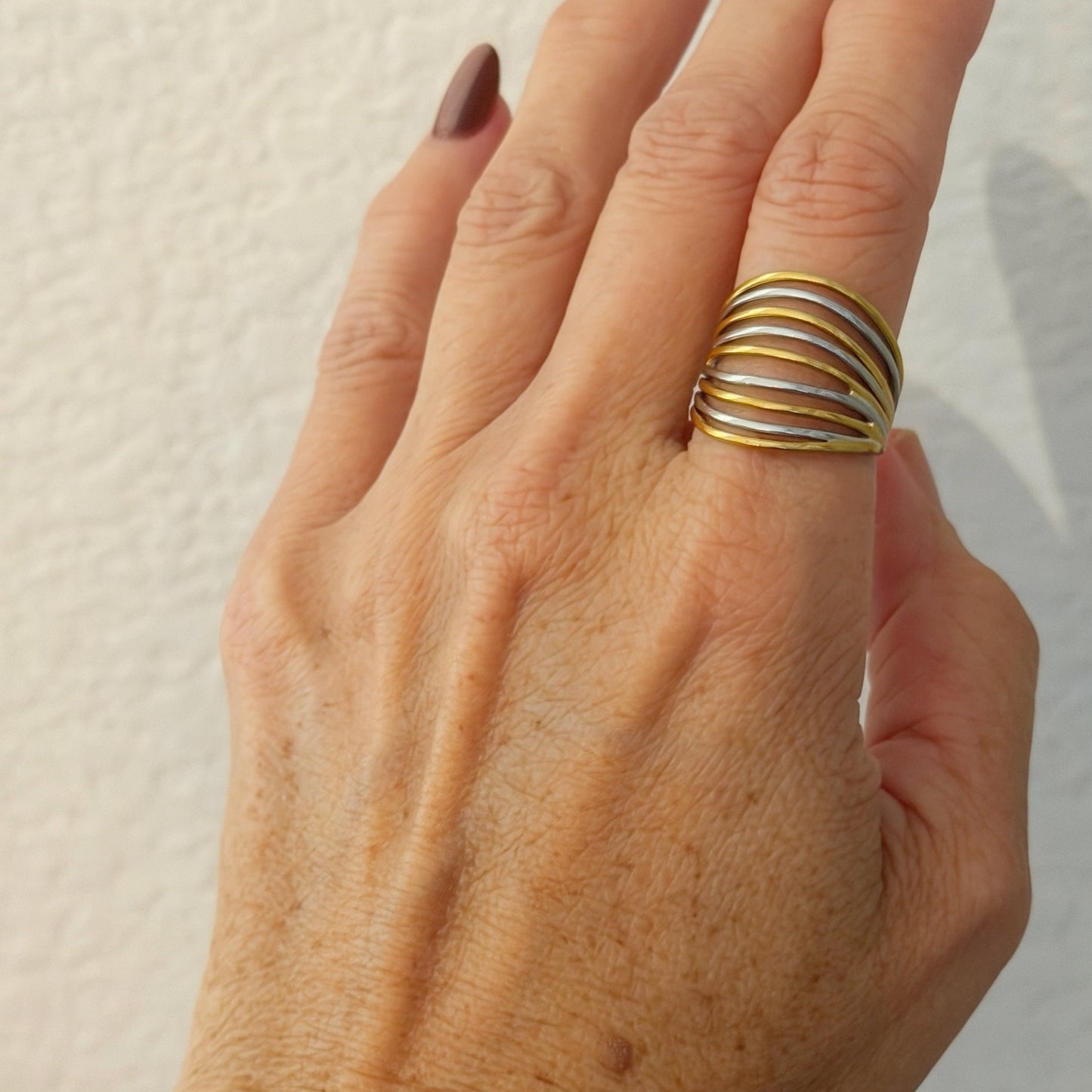 A close-up of a hand wearing a multi-band ring with alternating gold and silver-colored metallic bands, set against a textured off-white background.